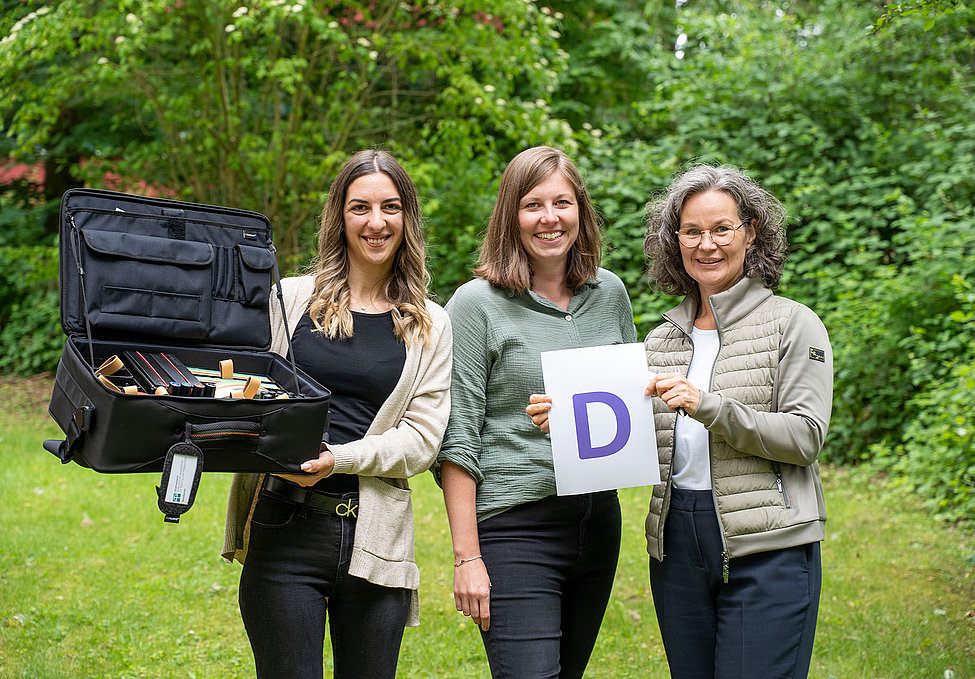 Drei Frauen halten Koffer mit Lernmaterialien und Blatt mit lila D