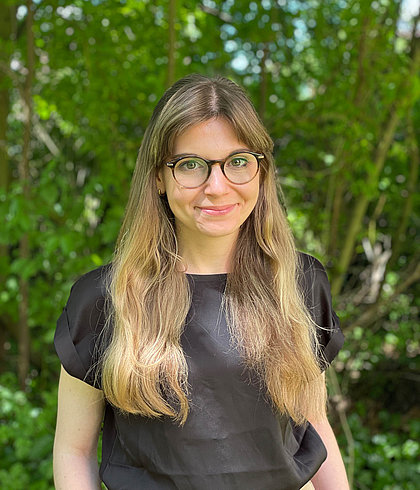 Porträt einer lächelnden Frau mit Brille und langen Haaren, schwarzes Oberteil vor dichtem grünen Laub.