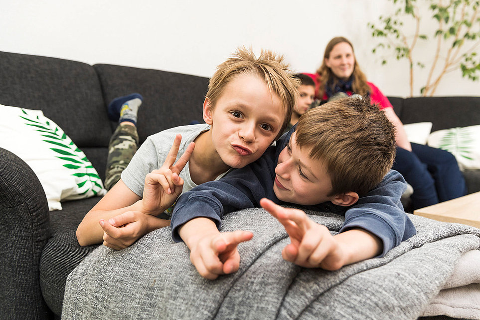 Zwei Jungen auf dem Sofa machen Friedenszeichen, Frau im Hintergrund