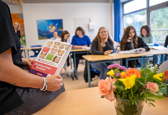 Lehrkraft hält Buch Erzieherinnen + Erzieher vor Schülern im Klassenraum