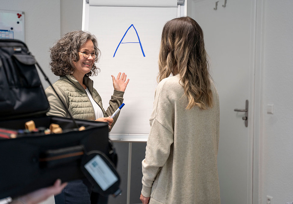 Trainerin erklärt Teilnehmerin vor Flipchart mit großem A