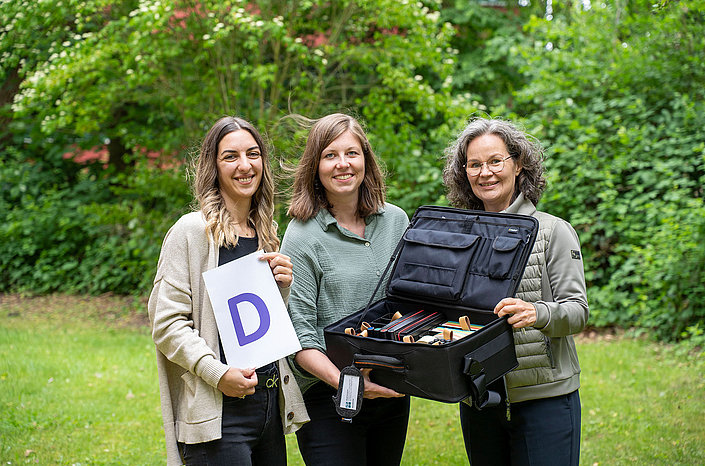 Drei Mitarbeitende halten eine offene Tasche mit Unterlagen und Schild mit D